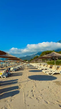 Saman gölgelerinin altında boş şezlongların, yukarıda mavi gökyüzünün ve dinlenmek ve huzurlu tatil anları için uzak mesafede dağların olduğu sakin bir sahil hayal edin.