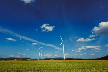 Bereketli yeşil alanlardaki yüksek rüzgar türbinlerinin çarpıcı görüntüsü, canlı mavi bulutlarla dolu gökyüzüne karşı yenilenebilir enerji ve doğa arasındaki uyumu gösteriyor.