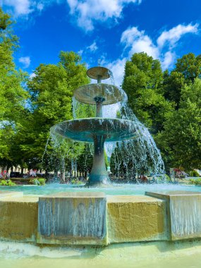 Güzel bir parkta, yeşil ve mavi gökyüzü ile çevrili zarif bir çeşme serinletici sular, günlük hayattan uzaklaşmak için ideal sakin bir atmosfer yaratıyor.