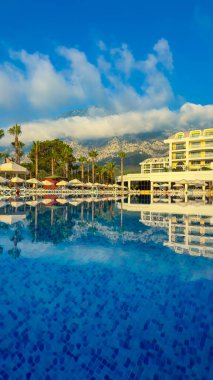 Huzurlu bir havuza yansıyan lüks bir otel. Parlak mavi bir gökyüzünün altında ve arka planda dağlar. Rahatlamak isteyen gezginler için mükemmel..