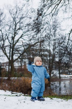 Mavi bir kar elbisesi içindeki neşeli bir çocuk, akmakta olan bir nehrin kenarındaki yumuşak karda oynar. Kışın keyfini kucaklayarak, macerayı davet eden güzel bir kar manzarasına karşı.