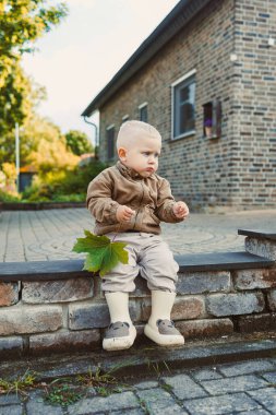 Neşeli bir çocuk taş basamaklarda oturur, canlı yeşil bir yaprak tutar, çocukluğun kaygısız maceracı ruhunu sembolize eder renkli, canlı doğal bir ortamda.