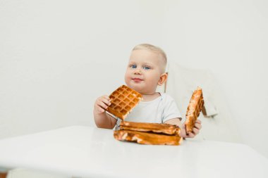 Sevimli küçük bebek, modern, temiz ve davetkar bir mutfak ortamında yüksek bir sandalyede otururken lezzetli, taze yapılmış waffle 'ları yerken mutlu bir şekilde atıştırıyor.