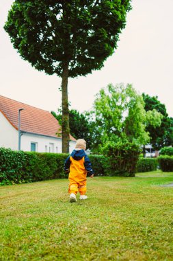 Parlak turuncu yağmurluk giyen neşeli bir çocuk, sakin bir banliyö mahallesinde büyüleyici, bulutlu bir gökyüzünün altında, yemyeşil çimlerde mutlu bir şekilde yürüyor.