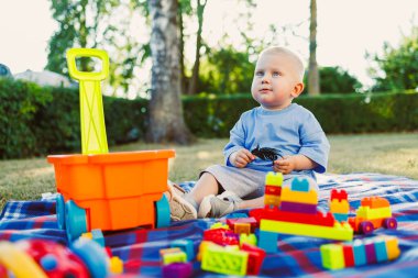 Sevimli bir bebek, güneşli ve güzel bir günde, açık havada, dışarıda oyun oynamaktan keyif alıyor. Etrafı renkli ve canlı oyuncaklarla çevrili, yemyeşil bir ortamda, rahat bir battaniyenin yanında.