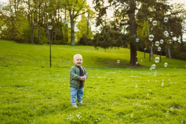 2 yaşında sabun köpükleriyle yeşil çimlerde koşan küçük bir çocuk. Küçük çocuk doğa yürüyüşünün tadını çıkarıyor. Çocuk şehir parkında eğleniyor..