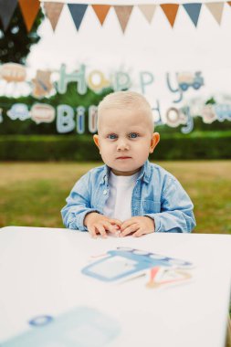 Sarışın, kıvırcık saçlı ve mavi gözlü çekici bir çocuk, doğum günlerini neşeli bir afişle dışarıda kutlamaya hazır bir masada oturuyor..