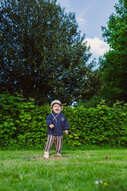 Kafasında panama şapkası olan güzel gülümseyen çocuk şehir parkında yeşil çimlerde koşuyor. Doğada iki yaşında bir çocuğun yürüyüşü. Çocuk doğanın güzelliğinin ve yazın güneşli bir günün tadını çıkarıyor..