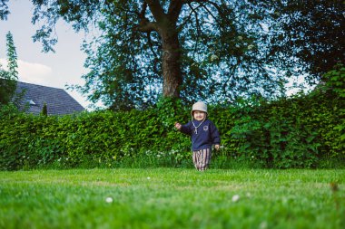 Kafasında panama şapkası olan güzel gülümseyen çocuk şehir parkında yeşil çimlerde koşuyor. Doğada iki yaşında bir çocuğun yürüyüşü. Çocuk doğanın güzelliğinin ve yazın güneşli bir günün tadını çıkarıyor..