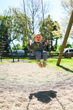 Kafasında Panama şapkası ve salıncakta sallanan açık renk ceketli sevimli 2 yaşında bir çocuk. Parktaki bir çocuk parkında salıncak. Bebek salıncakta sallanıyor. Şık çocuk kıyafetleri..