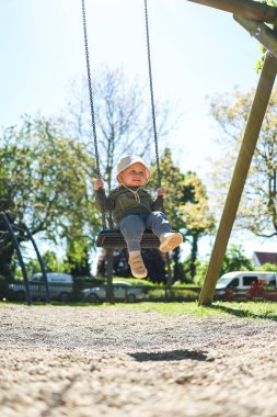 Kafasında Panama şapkası ve salıncakta sallanan açık renk ceketli sevimli 2 yaşında bir çocuk. Parktaki bir çocuk parkında salıncak. Bebek salıncakta sallanıyor. Şık çocuk kıyafetleri..