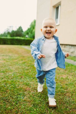 Neşeli bir çocuk yeşil çimenlerde koşuyor, kot ceket ve pantolon giyiyor. Çocuk açık havada güneşli bir ortamda eğlenirken saf mutluluğu gösterir..