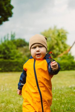 Parlak turuncu yağmurluk giymiş neşeli bir çocuk, yeşil bir tarlada neşeyle koşuyor, bulutlu bir gökyüzünün altında küçük bir çiçek tutuyor, tasasız çocukluk mutluluğunu somutlaştırıyor.