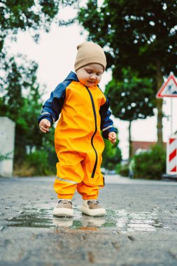 Parlak sarı yağmurluk ve rahat şapkalı neşeli bir çocuk, yağmurda oynarken çocukluk neşesinin özünü somutlaştıran çamurlu bir su birikintisine neşeyle sıçrıyor.