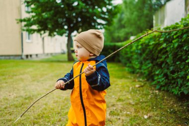 Parlak turuncu yağmurluk giymiş neşeli bir çocuk yeşil bir parkta sopayla oynar, hayal gücüyle dünyayı keşfederken neşe ve maceracı ruhu somutlaştırır