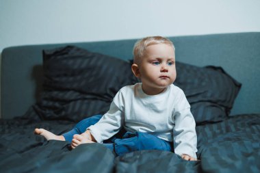 Yumuşak yastıkları ve sıcak battaniyeleri olan rahat bir yatakta, neşeli bir bebek oturur, sevgi ve merakla dolu sevgi dolu aile anları için mükemmel bir atmosfer yaratır.