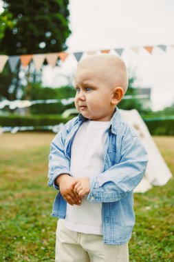 Şık bir kot ceket ve açık renk pantolon giymiş, şirin, küçük bir çocuk, neşeli bir açık havada duruyor güzel ve renkli bayrak ile süslenmiş.