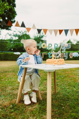 Sevimli genç bir çocuk neşeli bir şekilde bir masada oturuyor. Yanında güzel bir doğum günü pastası, canlı dekorlarla dolu açık hava ortamında, özel gününün tadını çıkarıyor.