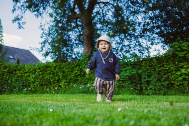 Mavi kapüşonlu neşeli genç bir çocuk, açık ve güneşli mavi gökyüzünün altında, çocukluğun kaygısız anlarının tadını çıkararak yemyeşil bir çimenlikte heyecanlı bir şekilde koşar.