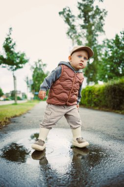 Yağmurlu bir günde, kahverengi yelek giymiş neşeli bir çocuk ve aynı kepçeyle bir su birikintisine sıçrıyor, çocukluk masumiyetinin özünü canlı yeşil bir manzarada yakalıyor.