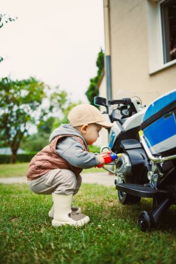 Canlı bir açık hava ortamında şapkalı ve botlu genç bir çocuk renkli bir oyuncak aracı hevesle inceliyor. Bu canlı anda meraklarını ve yaratıcılıklarını yansıtıyor.