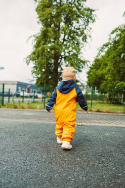 Parlak turuncu yağmurluk giymiş neşeli bir çocuk ıslak bir yolda yürüyor, açık havada yağmurun altında yemyeşil serpilmiş maceraların saf neşesini ve heyecanını sergiliyor