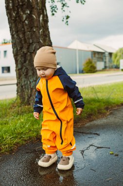Parlak turuncu yağmurluk ve rahat bir şapkayla neşeli bir çocuk açık havada su birikintilerinde neşeyle sıçrıyor, doğadaki çocukluğunun keyifli anlarını kucaklıyor.