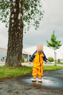 Parlak turuncu yağmurluk giyen neşeli bir çocuk, canlandırıcı yağmur duşu geçtikten sonra açık hava oyununun saf neşesini kucaklayarak su birikintisinde mutlulukla sıçrıyor.