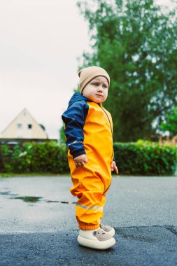 Canlı sarı yağmurluk giymiş şirin bir çocuk, ıslak bir zeminde duruyor. Etraftaki güzel doğal bir zemine karşı hem stil hem de neşe sergiliyor.