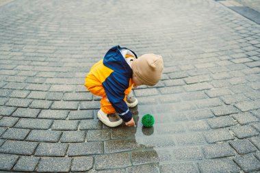 Sarı yağmurluk giymiş neşeli bir çocuk yeşil bir topla oynuyor, büyüleyici bir kaldırım taşı sokağındaki su birikintisinde neşeyle sıçrıyor, yağmurlu bir günün eğlencesini kucaklıyor