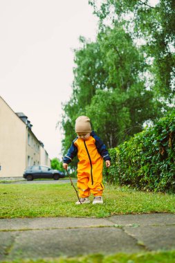 Parlak turuncu yağmurluk giymiş neşeli bir çocuk geniş yemyeşil çimenleri neşeyle keşfeder, elinde bir sopayla ve çocukluğun saf neşesini somutlaştırır.