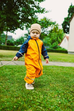 Sarı yağmurluk ve bronz şapkalı neşeli bir çocuk yemyeşil çimenlerde neşe içinde yürür bulutlu bir günde masumiyet ve neşe saçar açık hava macerasını kucaklarken