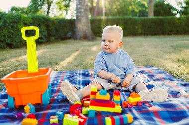 Neşeli küçük bir çocuk mutlu bir şekilde renkli bir piknik battaniyesinde oturur, neşeli bir şekilde açık havada güneşli bir yerde, parlak bir oyuncak arabasının yanında oyun taşlarıyla oynar.