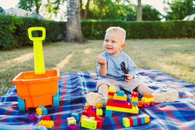Neşeli bir çocuk parktaki yumuşak çimenlerde piknik battaniyesinde renkli bloklarla oynuyor. Dışarıdaki eğlence ve yaratıcılık dolu oyun saatleri boyunca neşe saçıyor.