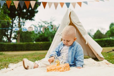 Güneşli bir günde neşeli bir bebek renkli bir battaniyeye oturur, güzel bir açık hava ortamında, canlı dekorasyonlarla çevrili doğum günü pastalarıyla mutlu olur.