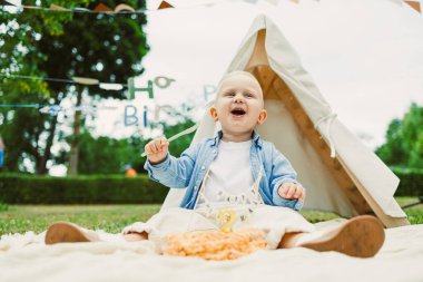 Neşeli bir bebek ilk doğum gününü renkli bir battaniyeyle pasta ve eğlence süslemeleriyle süslenmiş bir çadırın önünde kutlar. Saf mutluluğu simgeler.