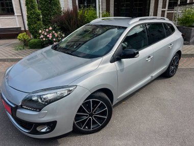 Siyah jantları olan şık, gümüş renkli bir hatchback, zarif ve canlı bir bahçenin önüne park edilmiştir. Modern tasarımı klasik zarafetle harmanlamıştır.