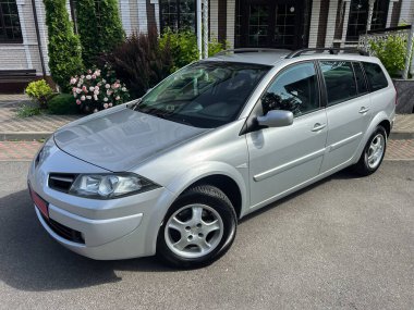 Gümüş renkli bir Renault Megane Sahnesi verimli yeşillikte zarif bir şekilde park edilir, güzel tasarımı ve aile maceraları ve eğlence seyahatleri için çok yönlülüğü vurgulanır.