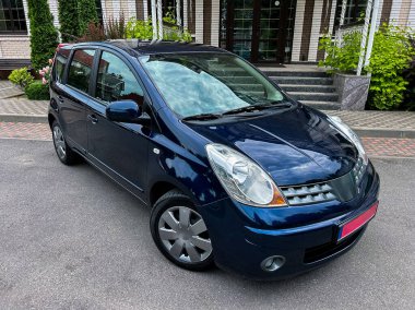 Canlı yeşilliklerle çevrili, modern bir binanın önünde zarif, koyu mavi bir araba, çekici tasarımı ve şehir cazibesini sergiliyor.