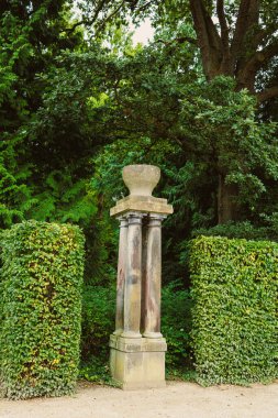 Zarif bir şekilde yaşlanmış yıpranmış bir taş sütun, budanmış çalıların arasında duruyor, huzurlu bir bahçe manzarasına bir göz atmak için huzurlu bir yansıma ve takdir için