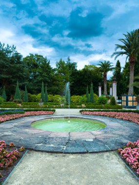 Sersemletici bir çeşme, canlı çiçekler ve kabarık bulutlu bir gökyüzü altında yemyeşil, huzurlu doğaya mükemmel bir kaçış için.