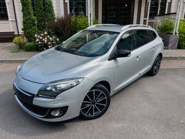 Parlak siyah alaşımlı tekerleri olan şık, gümüş renkli bir hatchback, canlı bitkilerle çevrili modern bir evin önüne park edilmiş.