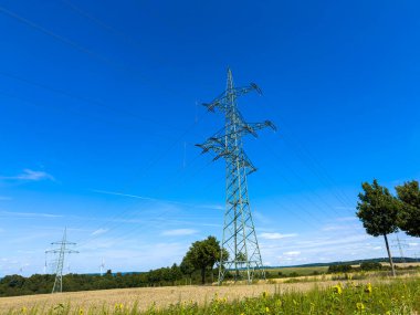 Yeşil alanlar ve ağaçlarla çevrili mavi gökyüzüne karşı yüksek voltajlı elektrik hatlarının resimsel görüntüsü modern hayatlarımızı birleştiren hayati enerjiyi sembolize ediyor.