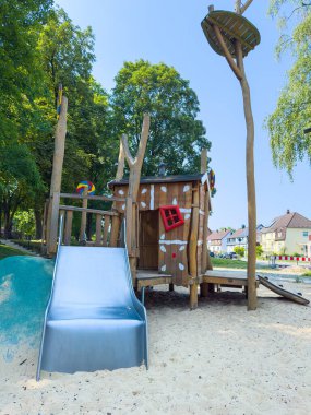 Tahta oyun evi ve parlak gümüş slaytlı, canlı ağaçlar ve kumlu zemin ile çevrili büyüleyici bir oyun alanı, çocuklar için eğlence ve oyun zamanı için ideal.