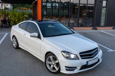 El değmemiş beyaz bir MercedesBenz coupe canlı bir şehir ortamında şık ve şık tasarımını arka plandaki şık camdan bir binaya karşı sergiliyor.