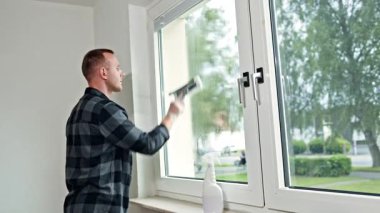 Genç erkek temizlikçi evin camlarını yıkıyor. Adam camları paspasla siliyor. Ev temizliği.