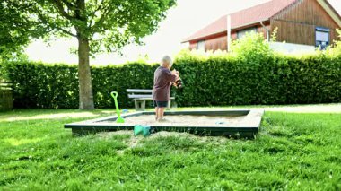 Kum havuzunda oynayan tatlı sarışın çocuk. Şortlu ve tişörtlü çocuk parkta kumla oynuyor. Yazın kum kazan bir çocuk. Çocuk gelişimi ve bağımsız oyun.