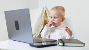 2 yaşında bir çocuk evde bir masada oturuyor ve bir dizüstü bilgisayara bakıyor. Bir çocuk taşınabilir bir bilgisayarda çizgi film izliyor. Modern bir çocuk ve bilgisayar..