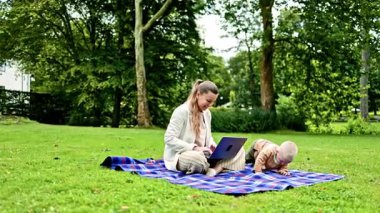 Parkta çimlerin üzerinde oturan ve dizüstü bilgisayarla çalışan çocuklu genç bir anne. Genç bir anne için bilgisayarla uzaktan kumanda. Anne ve oğlu parkta dinlenirken video izliyorlar.