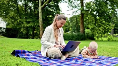 Parkta çimlerin üzerinde oturan ve dizüstü bilgisayarla çalışan çocuklu genç bir anne. Genç bir anne için bilgisayarla uzaktan kumanda. Anne ve oğlu parkta dinlenirken video izliyorlar.
